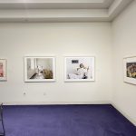 four works of art are hung in a line to the corner of a gallery. Royal Purple carpet lines the room. Two images are photographs of two girls waiting in an amtrak station and a man smoking in a hospital room. The two outer works of art are serigraphs