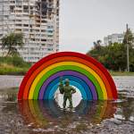 toy soldier stands infront of a toy rainbow in a puddle with a war torn city in background