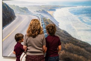 woman stands between two boys, all three gaze up at a landscape painting by artist Woody Gwyn with a highway curves around grassy mountains and the ocean on the right