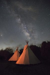 two teepees stand in an orange glow from a bonfire just out of frameagainst a night sky filled with stars and the milky way