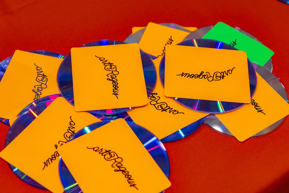 Compact Discs with a bright orange sticker that says "artRageous" are scattered across a bright red table